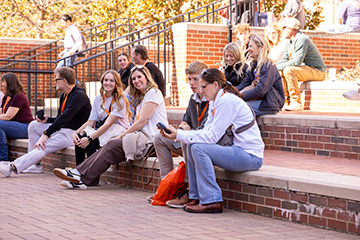 A group of college-age students sits together on brick steps along an outdoor campus plaza, casually talking and relaxing in a social setting during a campus visit or student event.