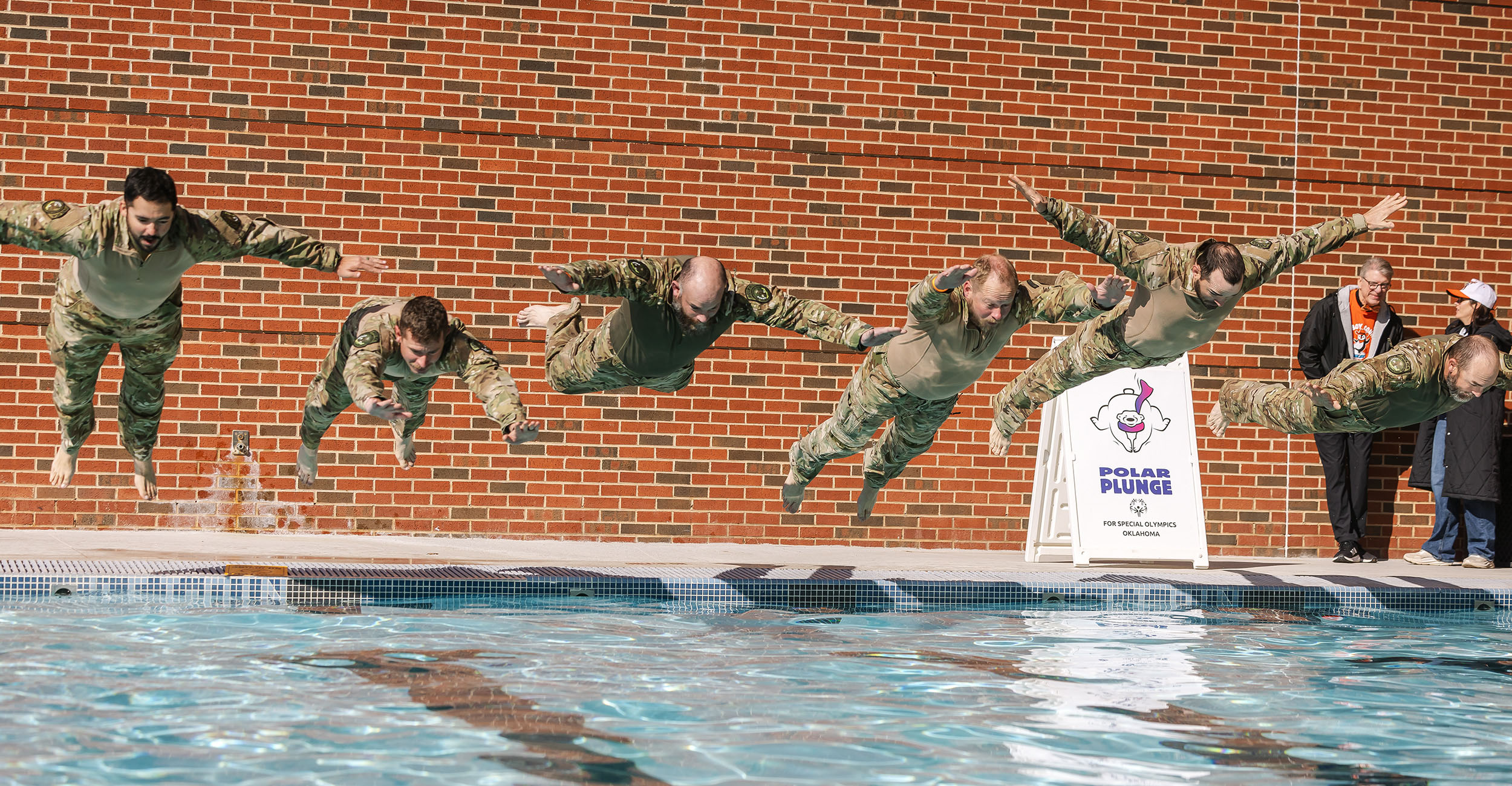 A group of people in camouflage uniforms leap simultaneously into a swimming pool during a Polar Plunge event, with a brick wall and event sign in the background.
