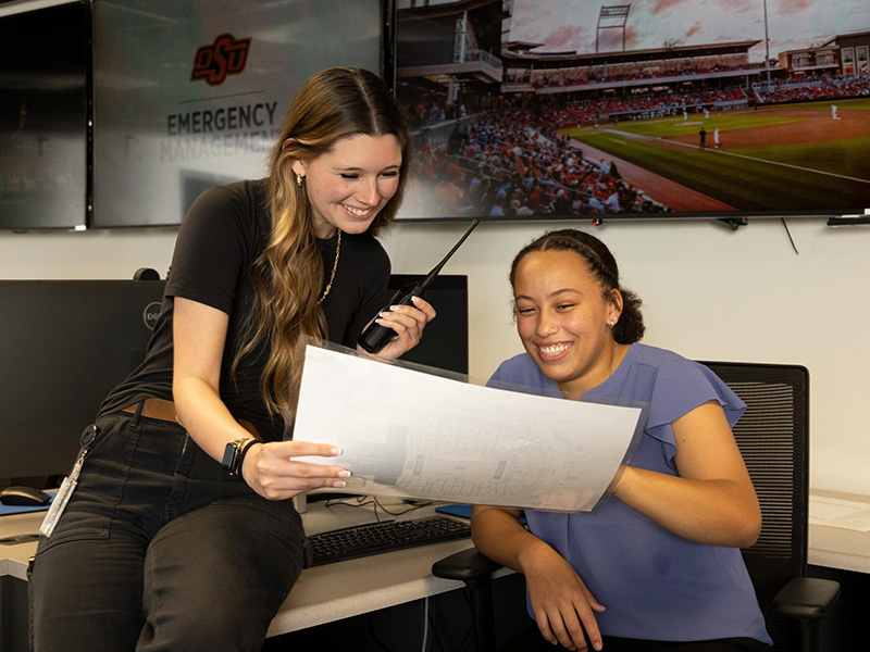  Two students sit at a workstation in an emergency management operations center reviewing a printed document together, with one holding a handheld radio and monitors in the background displaying OSU Emergency Management and a baseball game.