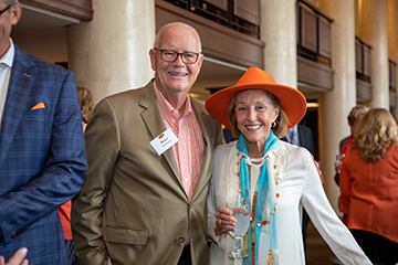 Two people pose together at an indoor event space, one wearing glasses and a name badge that reads “Bruce Benbrook” and the other wearing a bright orange hat, with columns and attendees visible in the background.