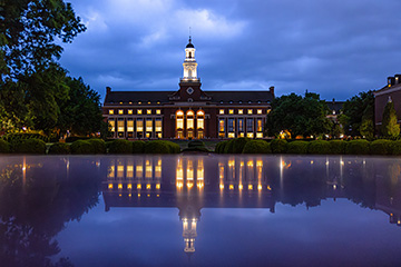 Oklahoma State University’s Edmon Low Library illuminated at dusk, with its tower and glowing windows reflected in the time capsule on Library Lawn beneath a cloudy blue sky.