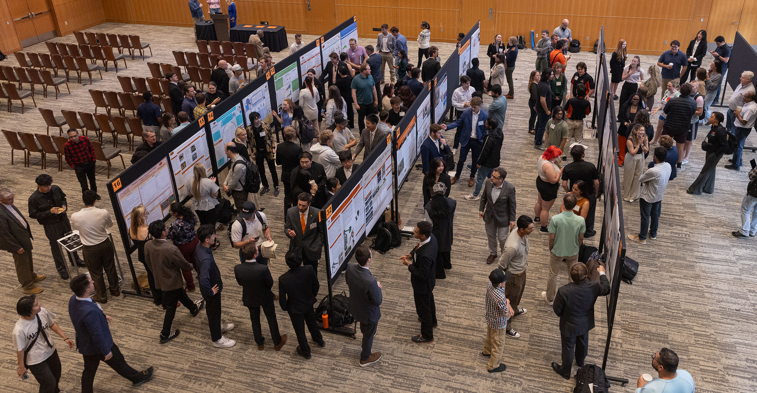 High-angle view of a large indoor hall at Oklahoma State University where dozens of undergraduate students stand beside rows of research posters, presenting and discussing their work with attendees.