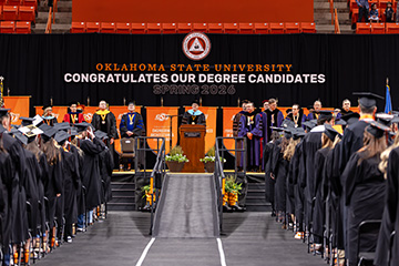 Wide-angle color photograph of an Oklahoma State University commencement ceremony with graduates in caps and gowns facing a stage where faculty in academic regalia stand beneath a banner reading “Congratulations Our Degree Candidates Spring