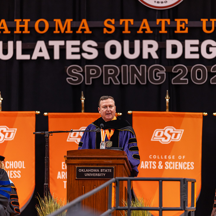 Color photograph of a person in academic regalia speaking at an Oklahoma State University podium on a stage with faculty seated behind and a banner reading “Congratulations Our Degree Candidates Spring 2026.”
