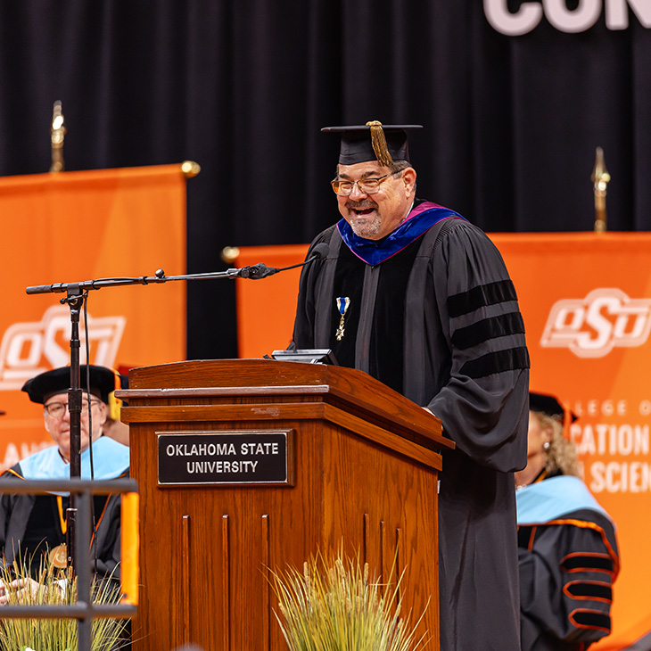 Color photograph of a person in academic regalia speaking at an Oklahoma State University podium during a commencement ceremony, with other faculty seated on stage behind.