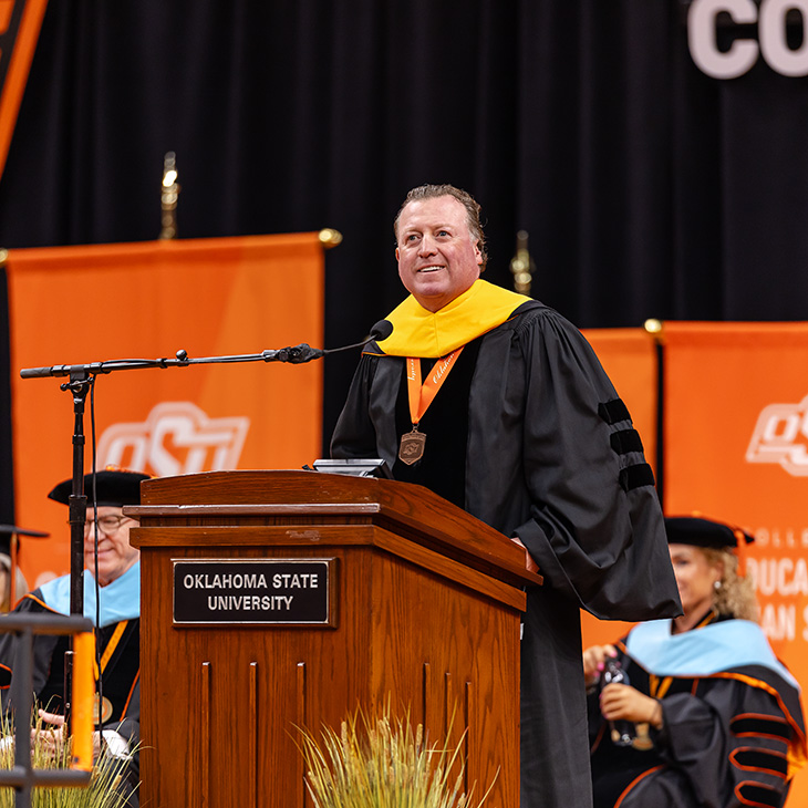Color photograph of a person in academic regalia with a gold hood speaking at an Oklahoma State University podium during a commencement ceremony, with faculty seated on stage behind and orange OSU banners in the background.