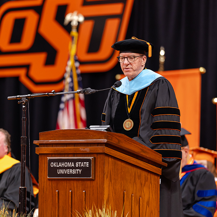 Color photograph of a person in academic regalia speaking at an Oklahoma State University podium during a commencement ceremony, with an OSU logo backdrop and faculty seated on stage behind.