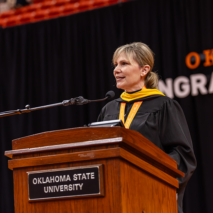 Color photograph of a person speaking at a podium labeled “Oklahoma State University,” wearing academic regalia with a gold hood, in front of a dark stage backdrop at a commencement ceremony.