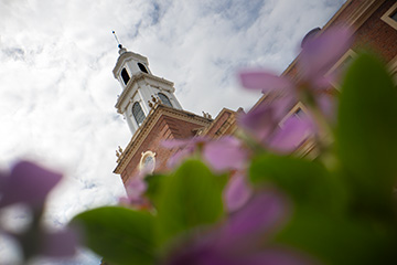 A low-angle view of Oklahoma State University’s Edmon Low Library tower rises above blurred purple flowers beneath a partly cloudy sky.