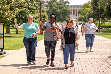 Participants walk along a campus sidewalk during an Oklahoma State University wellness walk, with several individuals gesturing and interacting as they move through a tree-lined outdoor setting.