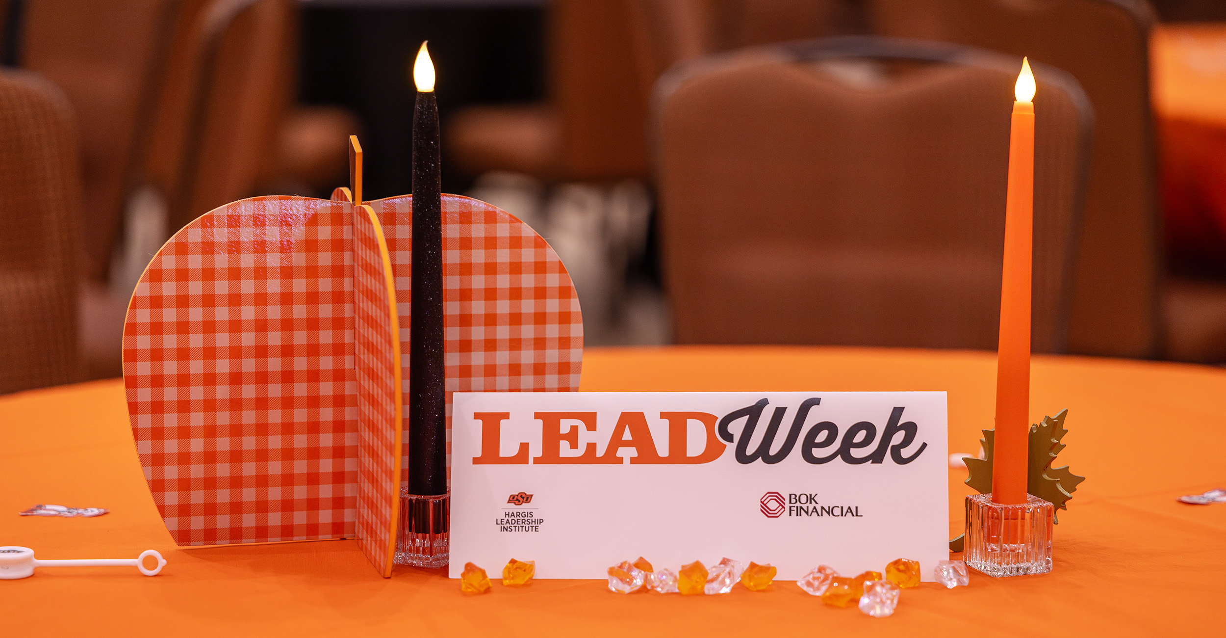 A LEAD Week event table centerpiece features decorative candles, a gingham-patterned paper pumpkin and a sign displaying the LEAD Week logo and sponsors on an orange tablecloth.