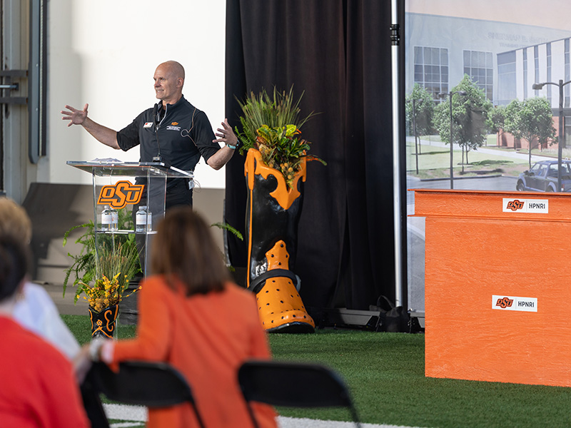 A speaker gestures while presenting from an OSU-branded podium during an event for the Boone Pickens Human Performance Innovation Complex, with attendees seated in the foreground and a rendering of the future facility displayed nearby.