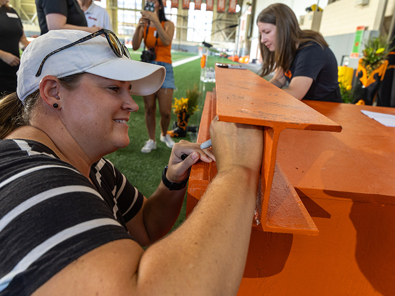 A participant signs an orange-painted steel beam during a ceremonial event inside an athletic facility, while others observe nearby before the beam’s installation in a future building project.