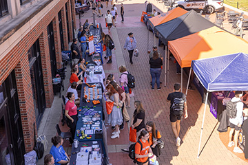 Students browse information tables and campus resource booths set up along an outdoor brick walkway during a campus fair, with vendors and attendees interacting beneath colorful event tents.
