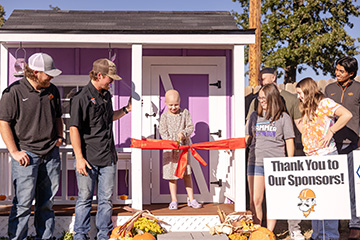 A child cuts an orange ribbon in front of a purple playhouse as Oklahoma State University students and others gather around for the dedication ceremony.
