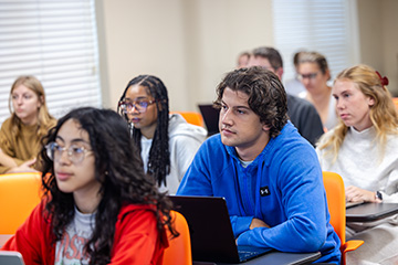 Students sit at desks with laptops while listening to a classroom presentation.