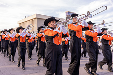 Oklahoma State University marching band members in orange-and-black uniforms and black cowboy hats play trombones while marching outdoors.