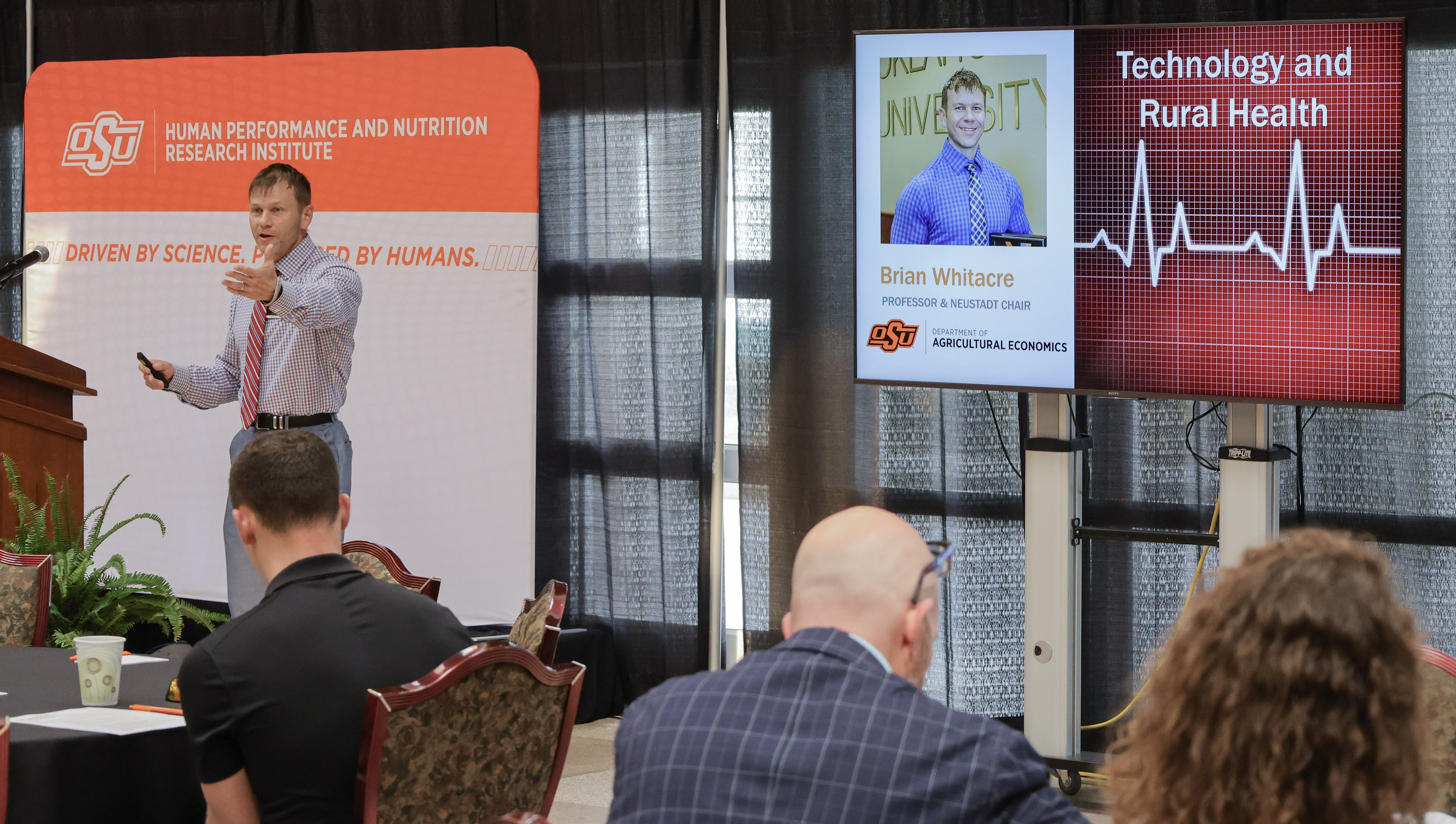 Dr. Brian Whitacre stands in the front of a room using a TV Screen to deliver a presentation at a research symposium. 
