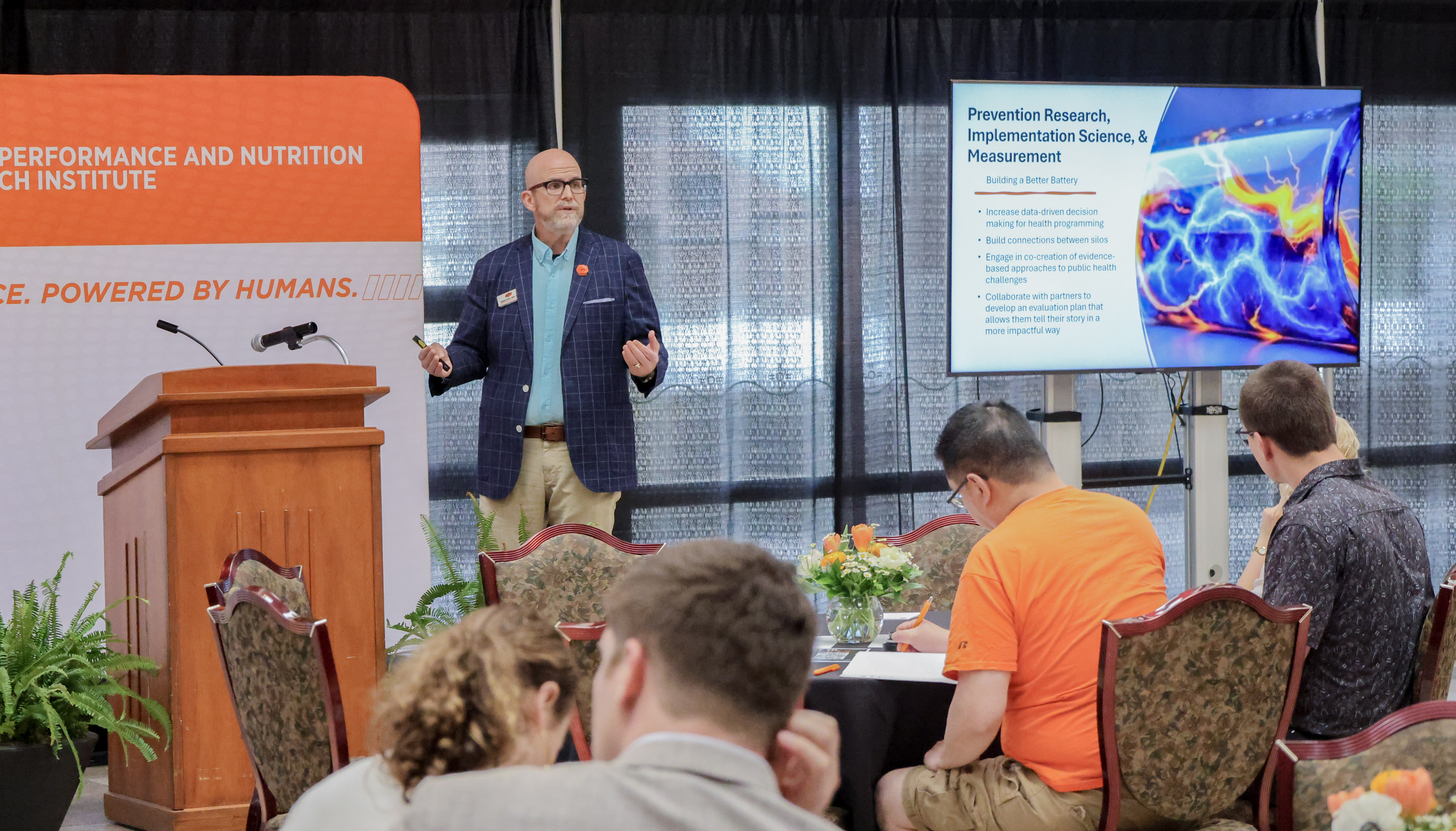 Chuck Lester stands in the front of a room using a TV Screen to deliver a presentation at a research symposium. 