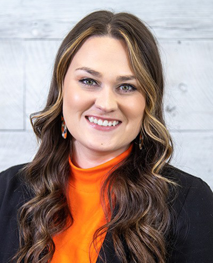 Professional head-and-shoulders portrait of a person wearing an orange turtleneck and black blazer against a light wood-paneled background.
