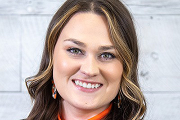 Professional head-and-shoulders portrait of a person wearing an orange turtleneck and black blazer against a light wood-paneled background.