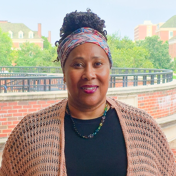 Portrait of a person standing outdoors in front of a brick terrace and campus buildings, wearing a patterned head wrap, dark top, necklace and knitted shawl.