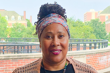 Portrait of a person standing outdoors in front of a brick terrace and campus buildings, wearing a patterned head wrap, dark top, necklace and knitted shawl.