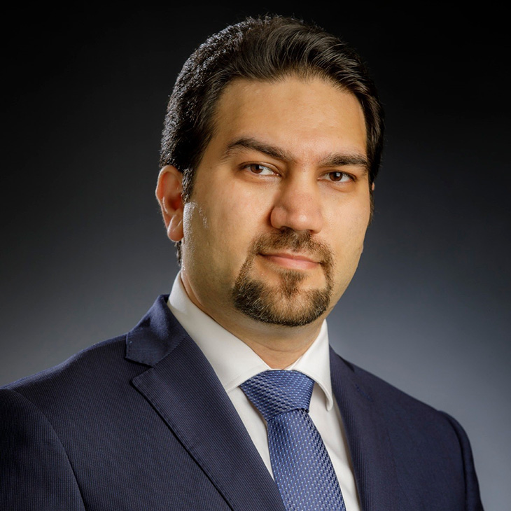 Formal head-and-shoulders portrait of a person wearing a dark suit, white shirt, and blue tie against a dark studio background.