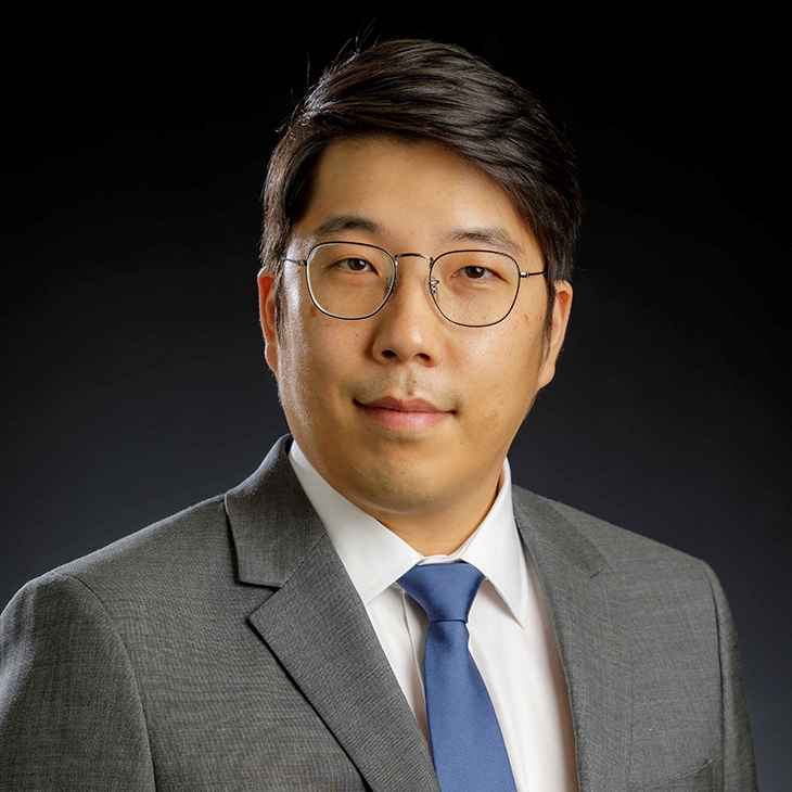 Formal head-and-shoulders portrait of a person wearing glasses, a gray suit, and a blue tie against a dark studio background.