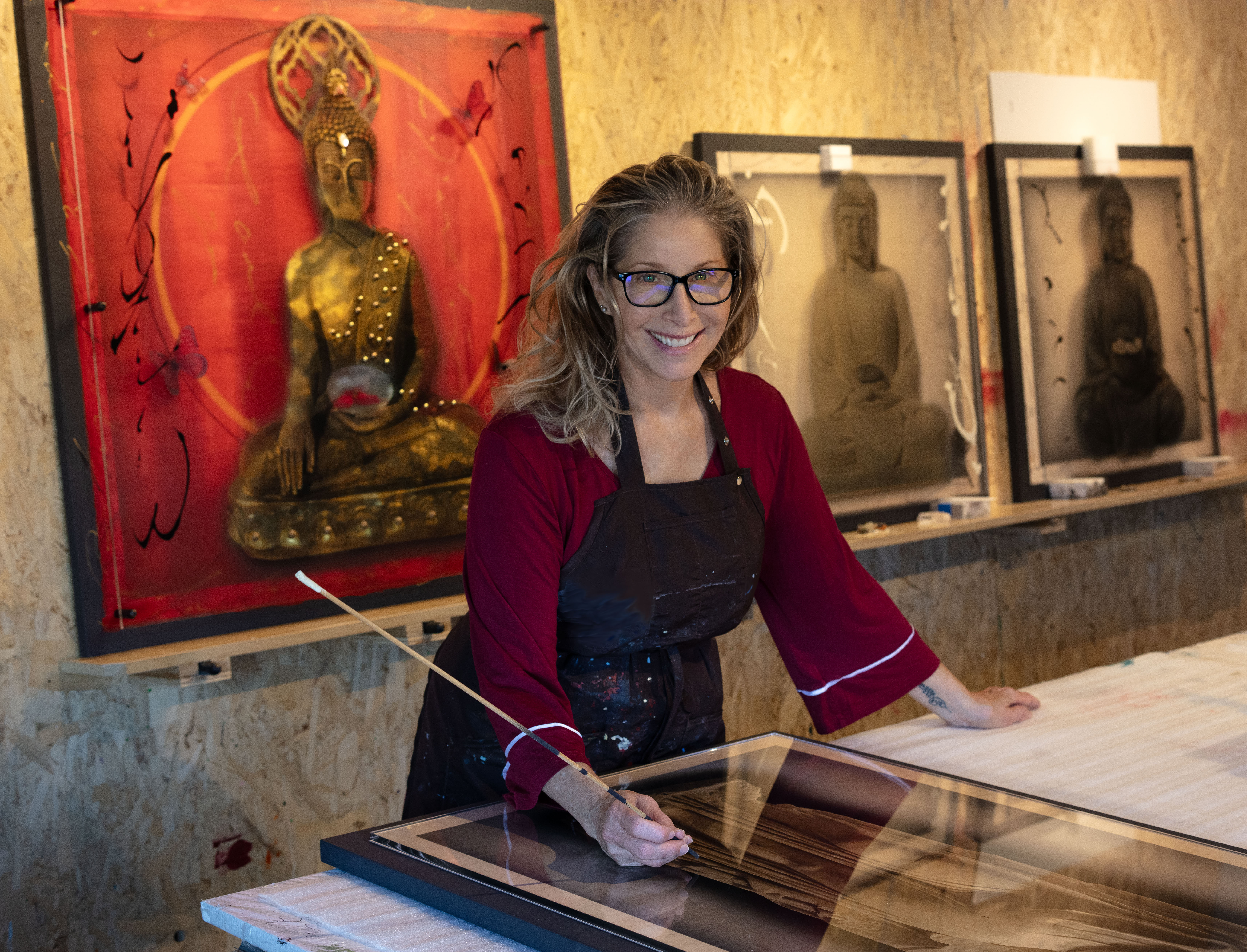 Person wearing glasses and an apron works on a mixed-media artwork in a studio, with framed Buddhist-inspired pieces displayed on the wall behind them, highlighting an artistic workspace.