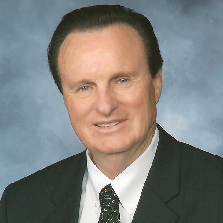 Formal head-and-shoulders portrait of a person in a dark suit and patterned tie against a blue studio background, facing the camera.