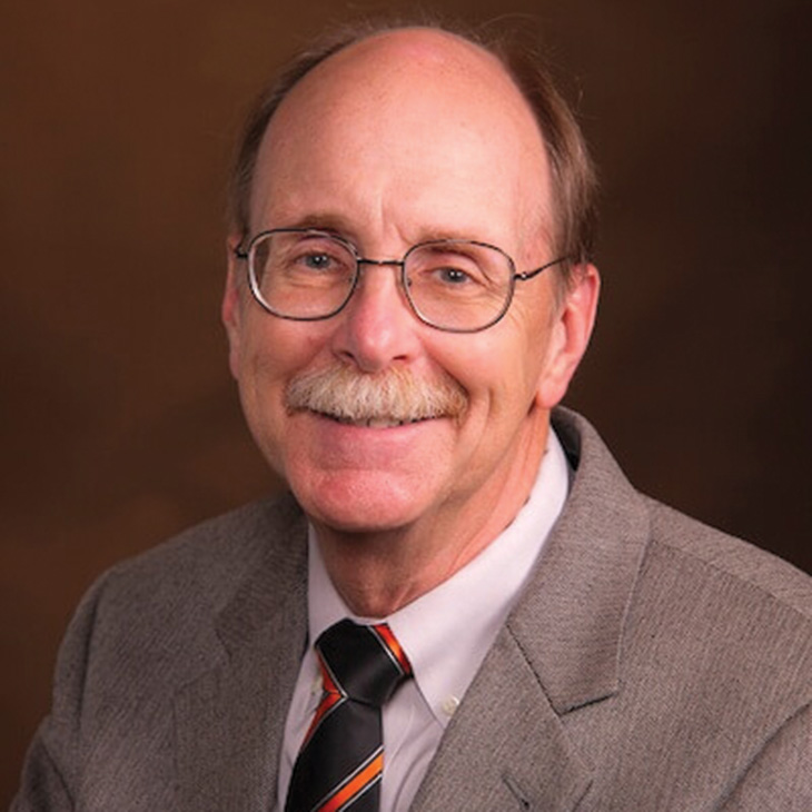 Head-and-shoulders portrait of a person wearing glasses, a gray suit jacket, and a striped tie, posed against a brown studio background.