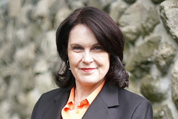 Professional portrait of a person with dark shoulder‑length hair wearing a black blazer over an orange shirt, posed against a textured stone wall background.