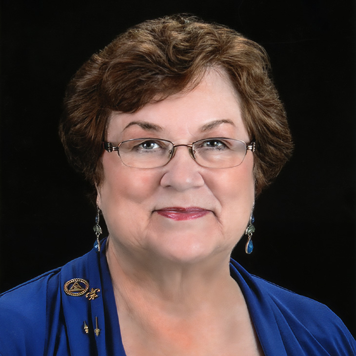 Formal headshot of a person wearing glasses and a blue jacket against a black background.