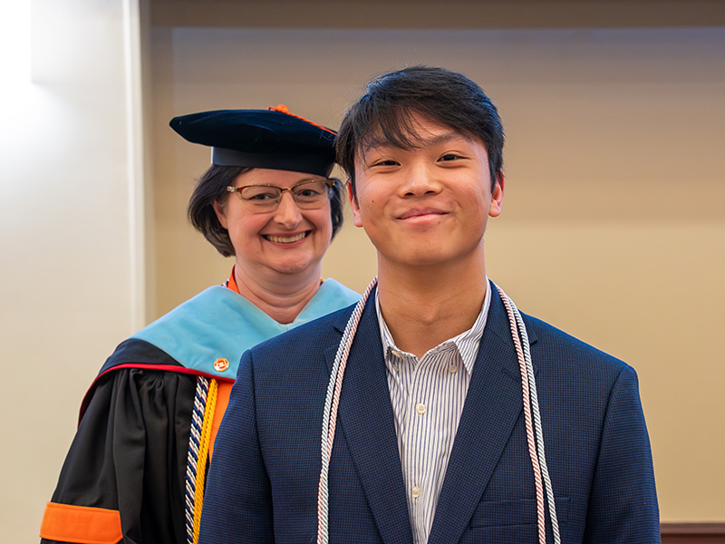 A student wearing honor cords stands for a recognition photo while an academic official in ceremonial regalia stands behind during an induction or awards ceremony.