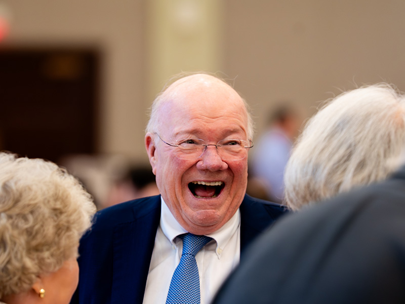 An attendee in a suit converses with others during a formal banquet or induction ceremony, captured in a candid moment among guests seated at tables.