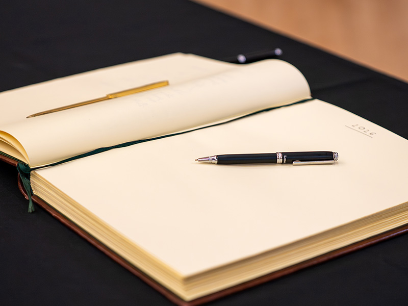 An open guest book rests on a table with two pens placed across its blank pages, ready for signatures or messages during a formal event.