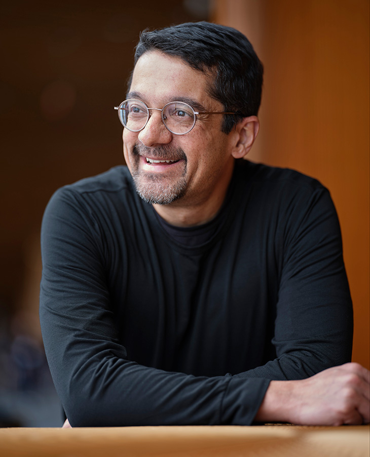 A person wearing glasses and a dark shirt sits at a table indoors, turned slightly to the side in a professional portrait-style photograph.