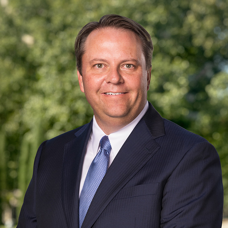 Formal headshot of a person wearing a dark suit and blue tie with blurred greenery in the background.