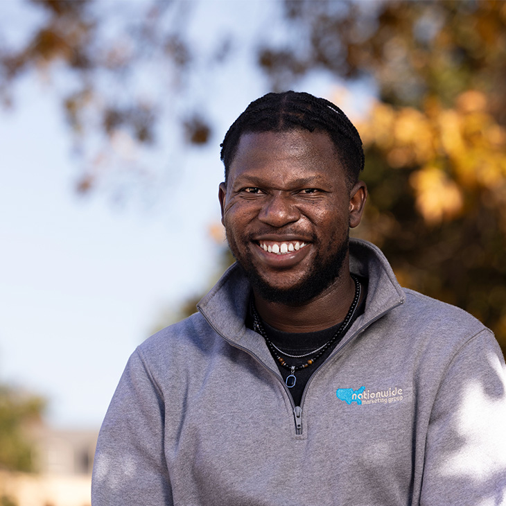 Adetokunbo Awonusi Person wearing a light gray quarter-zip sweatshirt with a blue “Nationwide Marketing Group” logo, layered necklaces, and sitting outdoors with autumn-colored trees in the background.