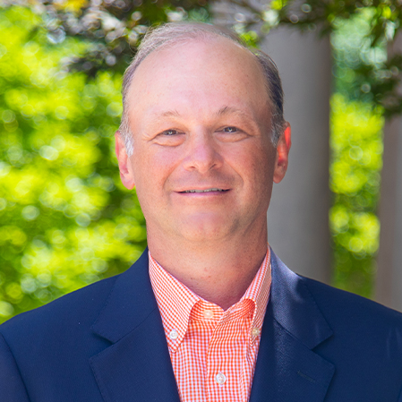 A man in a dark suit jacket and white collared shirt posed against a gray background