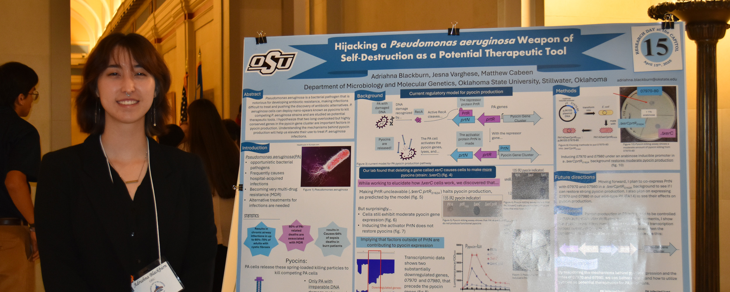 A student stands next to a research poster presentation at an academic event. The poster, displayed on an easel, features the Oklahoma State University logo and detailed charts, graphs, and text describing a scientific study. The student is smiling and wearing a conference badge and lanyard.