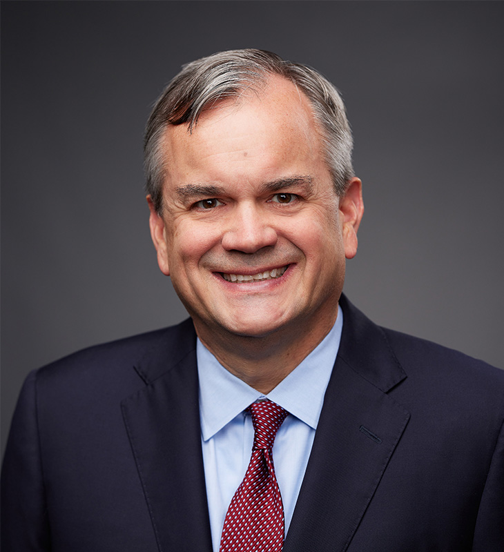 A man wearing a dark navy suit, light blue dress shirt, and red patterned tie smiles against a dark gray background.