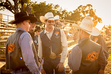 Several male students in cowboy hats and OSU branded vests talking