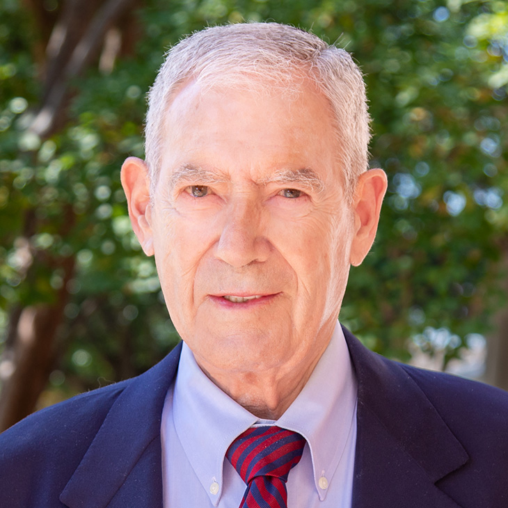 An older adult wearing a navy blazer, light blue dress shirt, and red‑and‑blue striped tie stands outdoors with green trees softly blurred in the background.