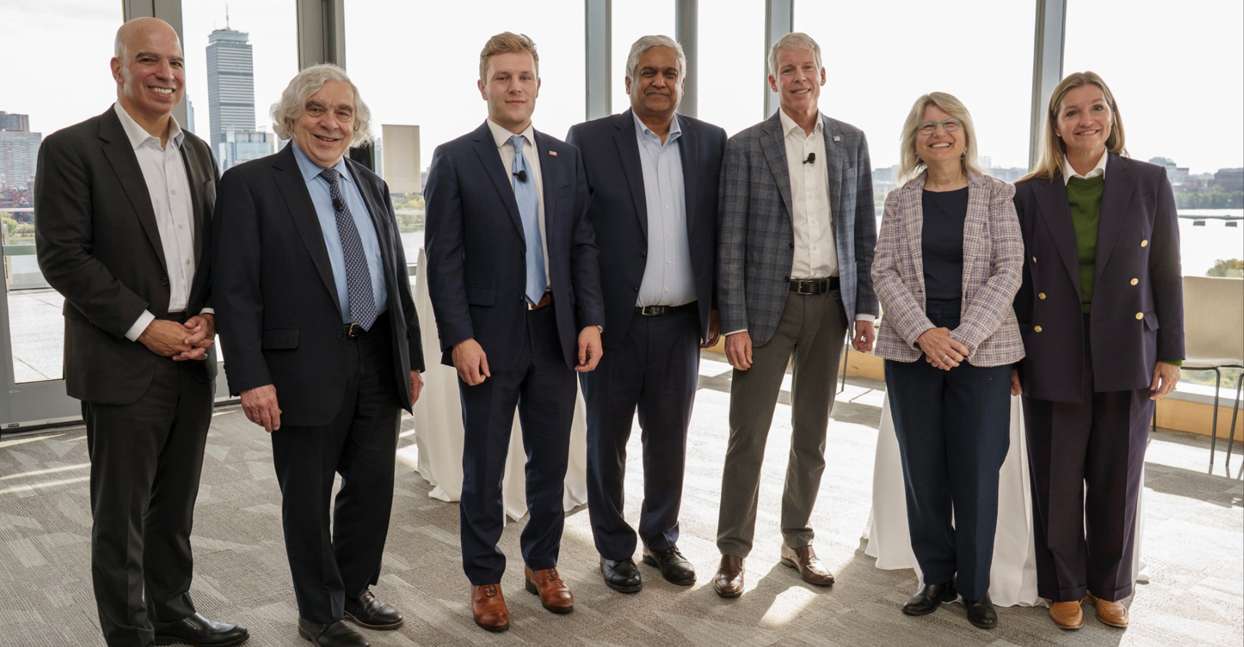 Group of six people standing indoors in front of large windows with a city skyline in the background. They are dressed in business attire, including suits, blazers and dress shoes.