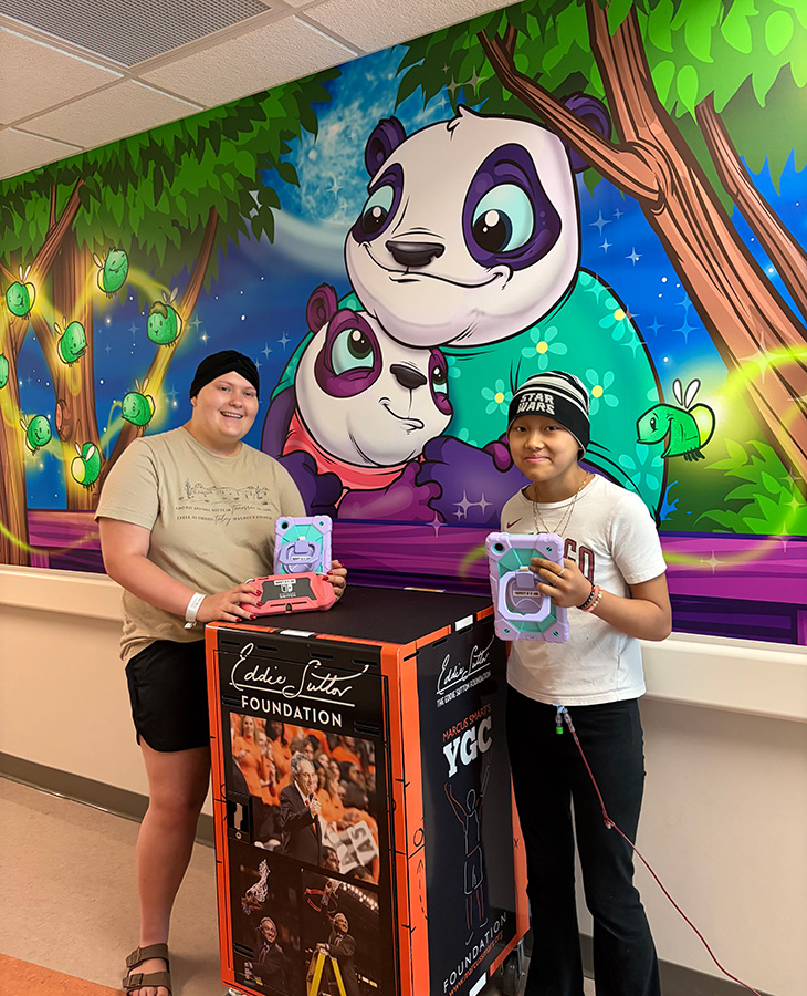 Two pediatric patients stand beside a medical equipment cart while holding tablet devices in a hospital hallway decorated with a colorful mural of cartoon pandas and forest animals.