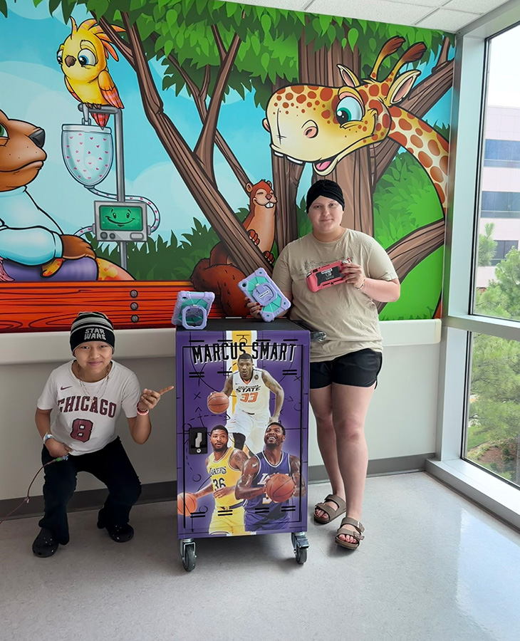 Two pediatric patients pose beside a mobile entertainment cart while holding gaming devices in a hospital hallway decorated with a colorful mural of cartoon animals, illustrating a program that provides recreational technology for patients.