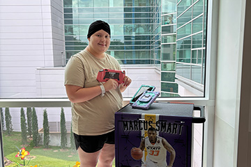 A patient stands beside a mobile entertainment cart while holding a handheld gaming device in a hospital corridor with large windows, illustrating a program that provides recreational technology for pediatric patients.