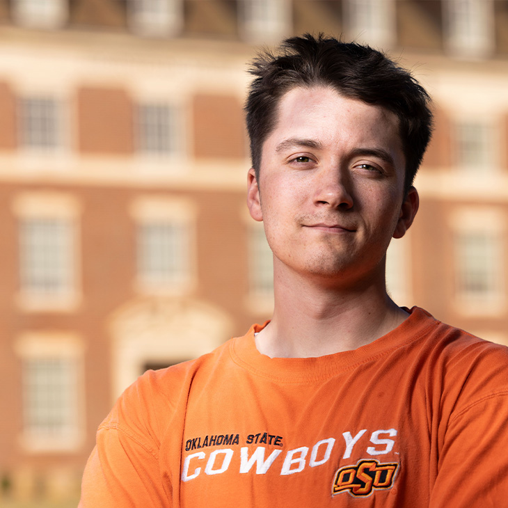 Ethan Kallweit Person wearing an orange Oklahoma State Cowboys T-shirt with OSU logo, standing outdoors in front of a brick building with large windows and architectural details.
